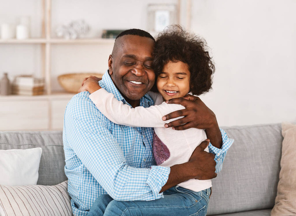 Залишайся вдома, будь щасливим. Афроамериканський дідусь обіймає свою прекрасну онучку у світлій кімнаті. - Фото, зображення
