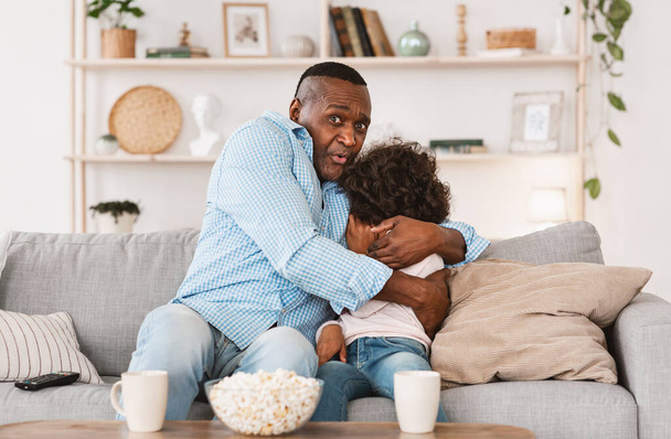 Entretenimiento familiar. El abuelo afroamericano abrazando a su nieta asustada mientras ve películas en casa
 - Foto, imagen