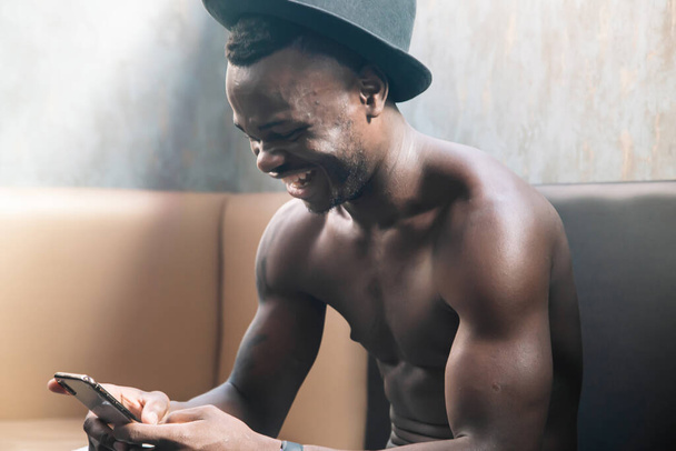 若い黒人男性は携帯電話を使用して革ソファーに座っている間笑います - 写真・画像