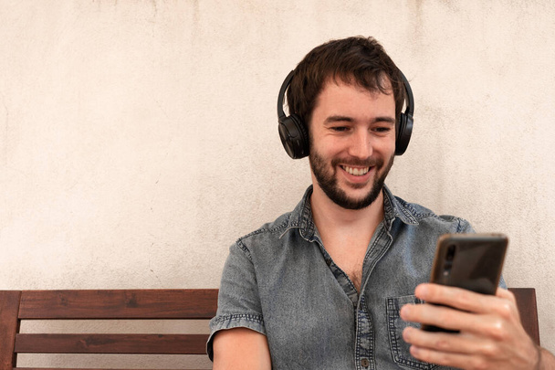 Pohledný chlapec s bluetooth sluchátka dívá na svůj mobilní telefon, a zároveň se usmívá pozitivně - Fotografie, Obrázek