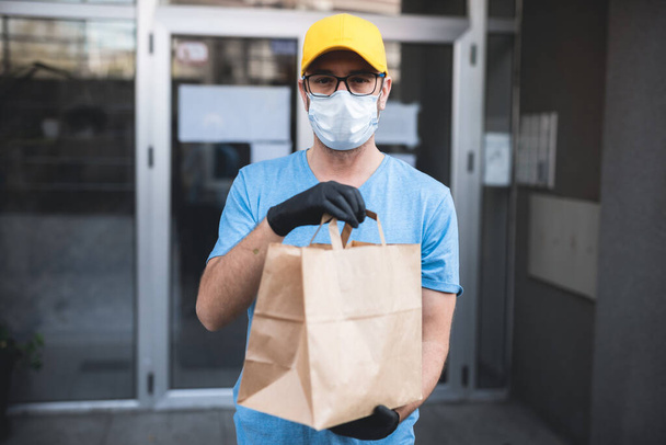 Доставщик в защитной маске и перчатках держит коробку / сумку с продуктами перед зданием
. - Фото, изображение