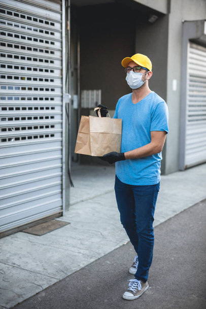 Koruyucu maskeli ve eldivenli teslimatçı. Bir binanın önünde poşet dolusu erzak tutuyor.. - Fotoğraf, Görsel