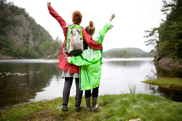 Portrait from behind mother and daughter standing by lake with arms raised - Photo, Image