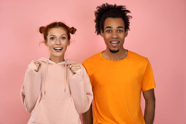 Photo of emotional ginger female and dark skinned male clench fists, exclaim and support favorite sports team, have overjoyed face expressions, dressed in casual wear, isolated on pink background - Fotó, kép