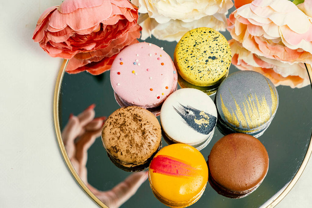 Woman's female hand reaches to sweet macaroons cookies in mirror reflection colorful modern gorgeous macaroons on mirror for celebration romantic party birthday anniversary pion flowers, top view - Fotografie, Obrázek