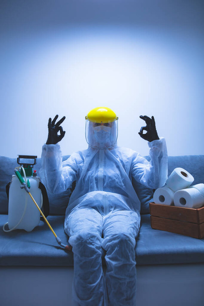 Person with toilet paper stock and protective antiviral mask, suit, helmet and chemical decontamination sprayer bottle in home isolation. - Фото, изображение