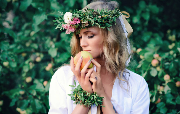 romantic girl with an apple and a wreath of flowers against the background of summer greenery - Photo, Image