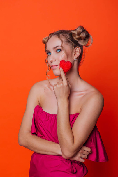 The girl in a red dress on a orange background in the studio. Blonde girl with two hair knots holding red heart on the stick. - Foto, Imagen
