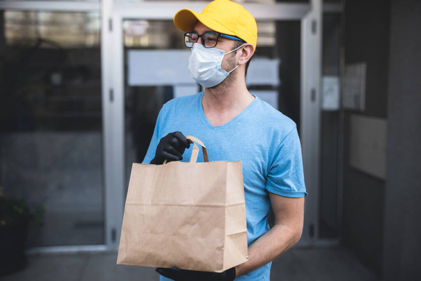 Dostawca z maską ochronną i rękawiczkami trzymający pudełko / torbę z zakupami przed budynkiem. - Zdjęcie, obraz