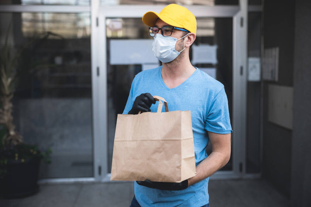 Хлопець з захисною маскою і рукавичками тримає коробку / сумку з продуктами перед будівлею
. - Фото, зображення