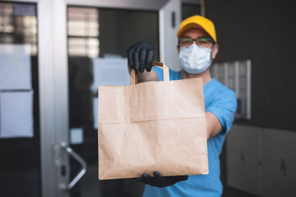 Хлопець з захисною маскою і рукавичками тримає коробку / сумку з продуктами перед будівлею
. - Фото, зображення