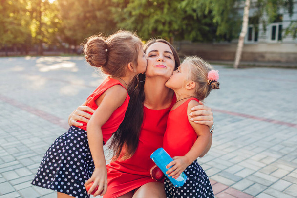 Dia da mãe. Filhas felizes beijando sua mãe depois de aulas ao ar livre escola primária com presente. Abraços familiares. As crianças adoram
 - Foto, Imagem
