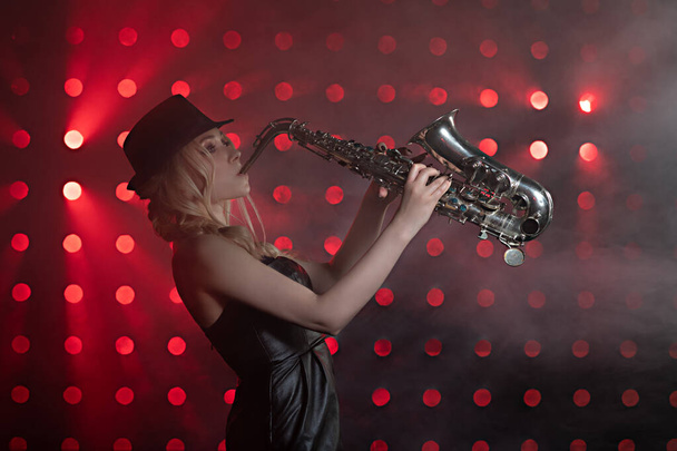 a girl plays a saxophone on a black and red glowing background in a black dress with heels - Foto, Bild
