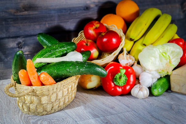 Vegetables and fruits on a graphite wooden background: in a box are tomatoes, cucumber, garlic, large red peppers, Iceberg lettuce, turnip onions, bananas, an apple, a basket with fresh carrots, cucumbers and fresh garlic is next to it. Still life, c - Photo, image