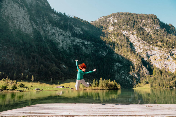 Молода жінка стрибає на пірсі біля озера під горами
 - Фото, зображення