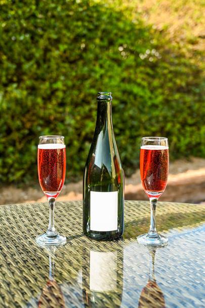 Zwei Flötengläser rosa Champagner und eine geöffnete Sektflasche mit einem leeren Etikett auf einer Glasplatte in einem Garten. Raum für Kopien. - Foto, Bild