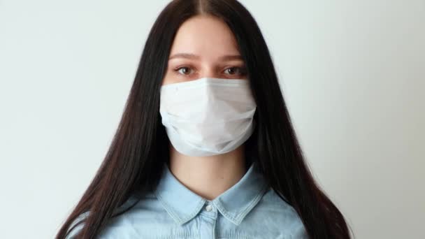 closeup of a young serious woman girl with long brown hair wearing a medical protective mask looking at the camera, concept of hygiene covid-19, coronavirus, on a white background, - Materiał filmowy, wideo
