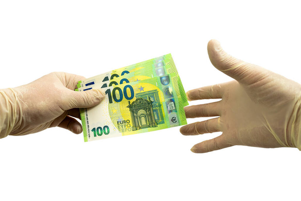 One hand in white protective gloves holds out a bundle of 100 Euro bills to the other hand in a white medical glove. Isolated white background. Virus protection concept - Foto, imagen