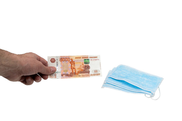 A hand buys a bill of five thousand five thousand Russian rubles several blue medical surgical masks. The concept of protection against the virus in Russia. Isolated background. - Photo, image