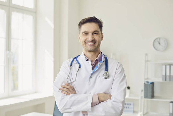 Positive doctor therapist in a white coat crossed arms standing in a white office of a medical clinic. - Photo, Image