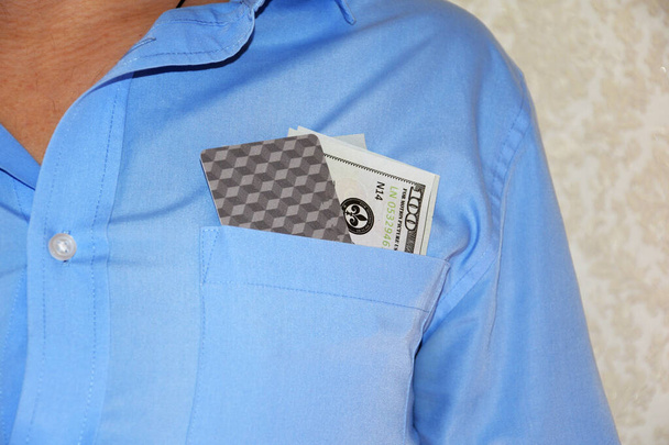 Στην τσέπη ενός άνδρα μπλε πουκάμισο είναι δολάρια και μια διεθνής κάρτα του συστήματος πληρωμών. - Φωτογραφία, εικόνα