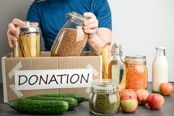 Donation box concept. Adult Caucasian man fills a cardboard box with food on a dark table. Voluntary assistance to those in need. - Photo, Image