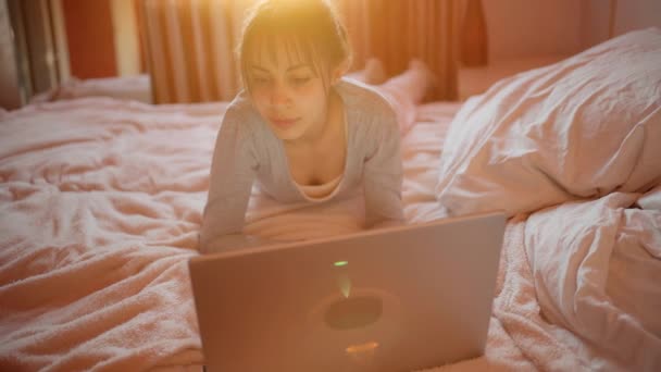 young woman in pajamas using laptop computer while relaxing in bed at home, sunlight in bedroom. - Séquence, vidéo