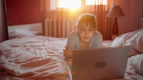 Young woman lying on bed at home and watching laptop computer, browsing on Internet. - Кадры, видео
