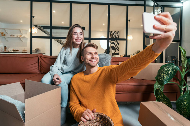 Feliz joven adulto novia y novio se mueven en un nuevo alojamiento juntos, sentado cerca de cajas de cartón, haciendo auto foto en el teléfono inteligente moderno
 - Foto, Imagen