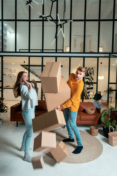 Relocation concept. Vertical photo of shocked young adult man and scared woman move in new flat together, dropping cardboard boxes, looking how package fall on floor - 写真・画像
