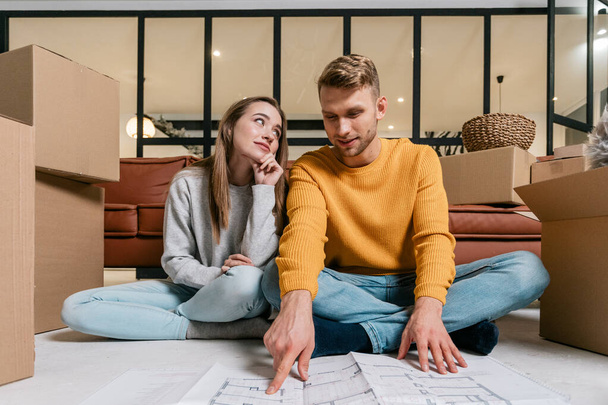 Low angle view of thoughtful and calm young adult man with woman move in rent accommodation together, sitting near blueprint, pointing on plan and discussion new interior in room - Photo, Image