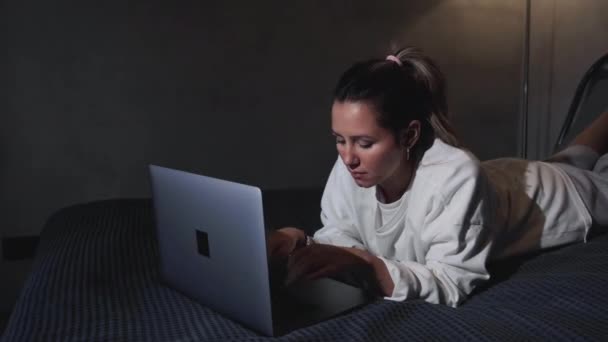 Woman have breakfast in modern hotel apartment. Window light portrait young girl in bathrobe lying on bed using laptop for work. Out of focus tray with meal, juice, plate, teapot on background - Footage, Video