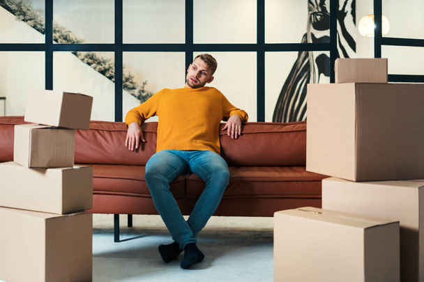 Измученный молодой человек переезжает в новую квартиру, сидит на диване, смотрит на коробки с вещами, отдыхает и готовит вещи
 - Фото, изображение