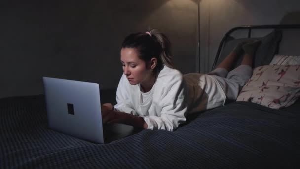 Portrait of girl lies in bed on her stomach, types on laptop keyboard, slow motion - Imágenes, Vídeo