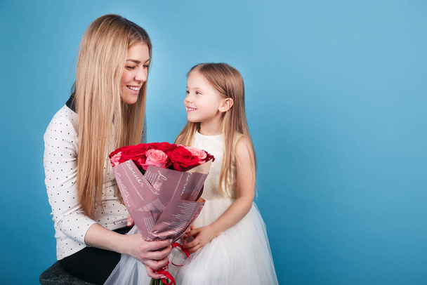 Erstaunlich fröhlich freudig charmante Mutter hält schöne Blumen Rosen und schaut auf ihre kleine süße Tochter auf blauem Hintergrund - Foto, Bild