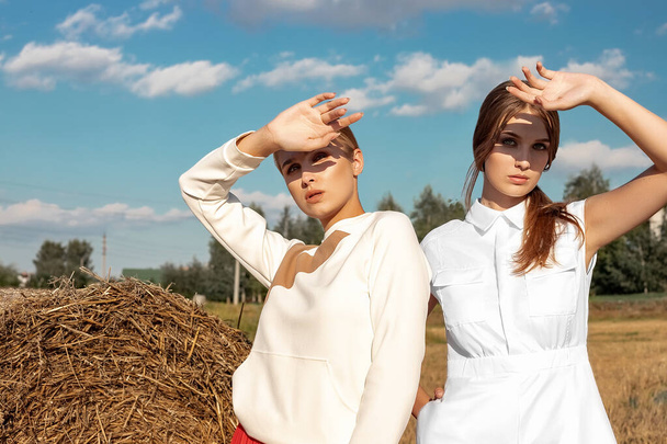Portrait of two fair-haired girls in fashionable and stylish clothes, against the background of a field and a stack of straw. On a sunny day in August. - Photo, image