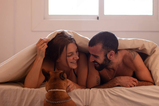 Happy couple lying on bed while a dog tries to go up with them - Photo, Image