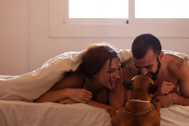 Glückliches Paar liegt auf dem Bett, während ein Hund versucht, mit ihnen nach oben zu gehen - Foto, Bild