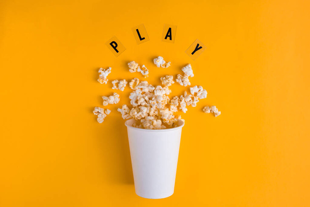 Paper cup with popcorn and text PLAY on yellow background. Flat lay banner, To go to the cinema. I like watching films. - Photo, Image