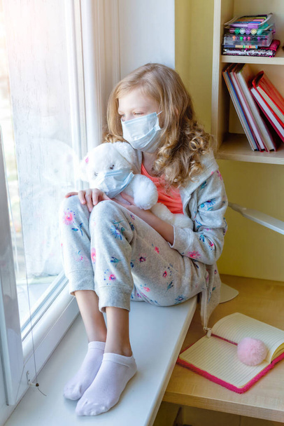 ragazza in maschera medica con orsacchiotto, guardando fuori dalla finestra, quarantena coronavirus. bambini malati
 - Foto, immagini