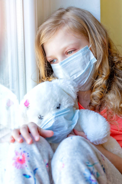 девушка в медицинской маске с плюшевым мишкой, выглядывает в окно, карантинный коронавирус. больные дети
 - Фото, изображение