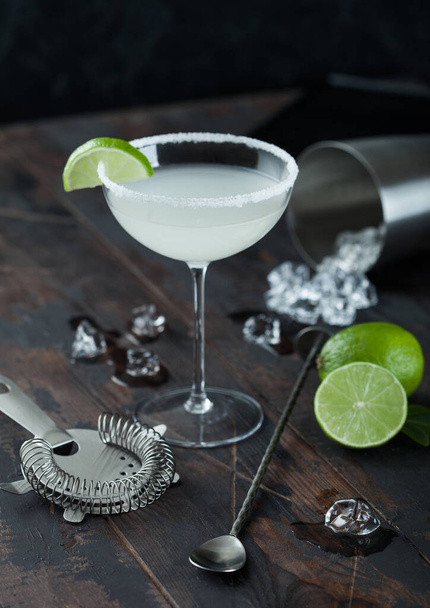 Glass of Margarita cocktail with fresh limes and bar spoon with strainer and ice cubes on wooden table background. Popular summer cocktail - Fotoğraf, Görsel