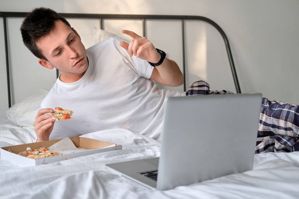 Young man in the white shirt and pajama watching video on laptop, cheering for the sports team lying on a bed and eating tasty pizza. Concept of food delivery. Staying home during quarantine. - 写真・画像