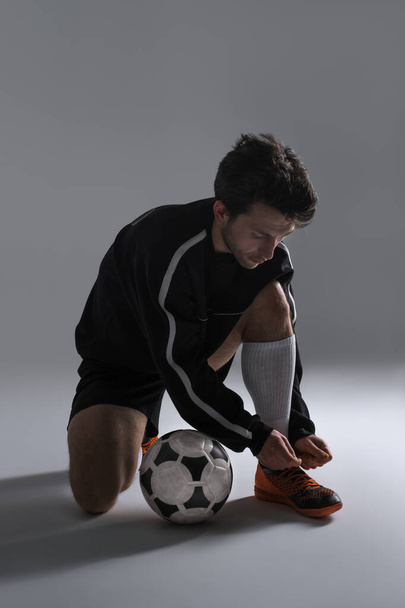 Jugador de fútbol en una ropa deportiva negra atando cordones de botas de fútbol aislados sobre fondo gris. Entrenamiento para la calificación de Copa de fútbol. Pelota de fútbol cerca de los pies de un jugador
. - Foto, Imagen