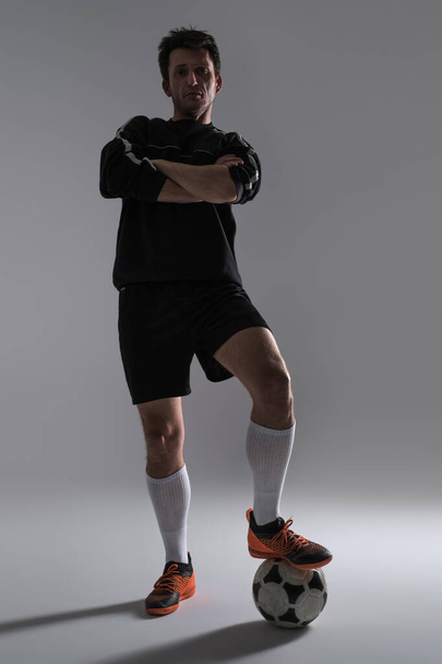 Selbstbewusster Fußballer in schwarzer Sportbekleidung, die Hände über dem Kopf verschränkt und isoliert vor grauem Hintergrund in die Kamera blickend. Training für die Fußball-WM-Qualifikation. Spieler hält Fuß auf einem Fußballball. - Foto, Bild