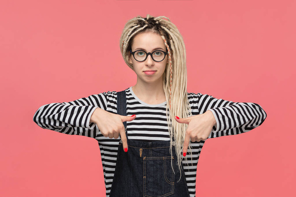 Schöne junge Mädchen mit Dreadlocks in einem gestreiften Hemd und Jeans-Jumpsuit, die mit zwei Fingern nach unten zeigen. Platz für Werbung - Foto, Bild