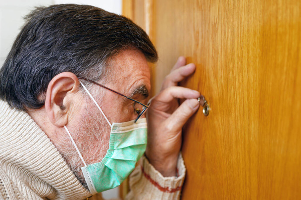  Covid-19 concept. Stay at home. Self-isolation to prevent the coronavirus pandemic. Senior man in protective green mask looks through the peephole. - Photo, Image