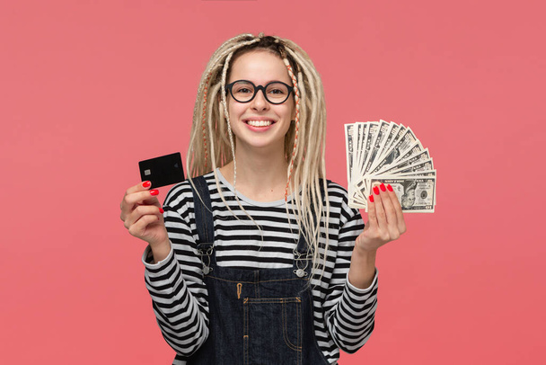 Excited teenager with dreadlocks in a striped shirt and jeans jumpsuit holding cash and bank card. Being rich - Photo, Image