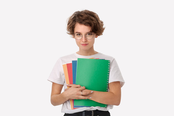 Smiling young female student in a white shirt and eyeglasses holding colorful folders isolated over white background. Concept of education - Photo, Image