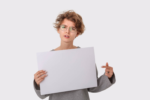テキストのためのコピースペースを持つ空の空白の紙板を保持し、指でそれを指す眼鏡を持つ懸念の若い女性。紙本著作物 - 写真・画像